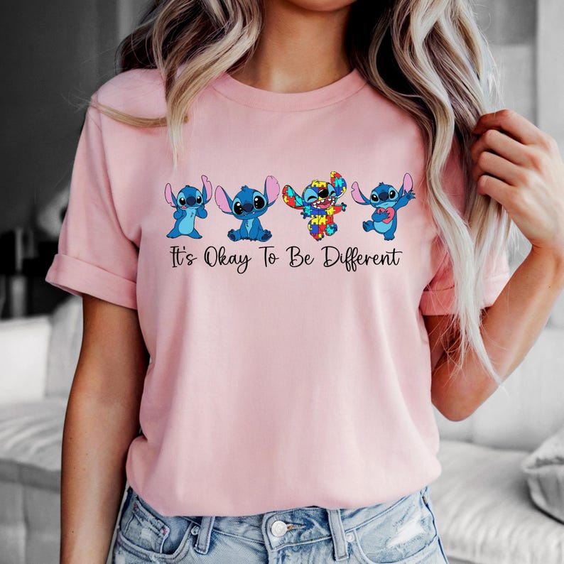 May include: Pink t-shirt featuring four cartoon Stitch characters in various poses. The text "It's Okay To Be Different" is printed below the characters in black. The shirt has short sleeves and a crew neck.