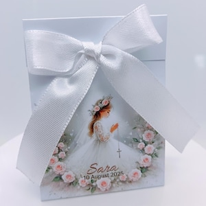 Puede incluir: Una caja de regalo blanca atada con una cinta de satén blanca. La caja presenta una ilustración de una niña con un vestido blanco, rodeada de rosas rosas, con el nombre "Sara" y la fecha "10 de agosto de 2025".