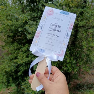 Peut inclure: Un éventail de mariage blanc avec un motif floral dans des tons de rose et de bleu. L'éventail a une poignée en bois et un ruban blanc. Le texte sur l'éventail indique "Veuillez vous joindre à nous pour célébrer le mariage de Sacha + Jamie le 20 août 2025, le dimanche à 12h00 123 Anywhere St, Any City, ST 12345 RSVP: 123-456-7890"