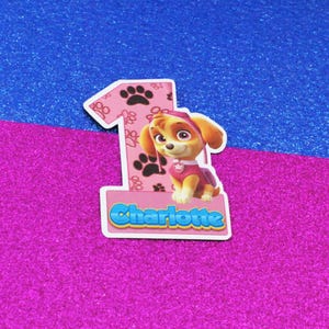 May include: A pink number one cutout with black paw prints and a cartoon puppy character. The puppy is wearing a pink outfit and a pink hat. The name "Charlotte" is written in blue letters.