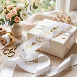 May include: A white satin ribbon wrapped around a square gift box, featuring gold foil text that reads Amelia & Sebastian and EST. 2024 with a laurel wreath icon. The ribbon is displayed alongside a pair of gold scissors and a small vase of flowers.