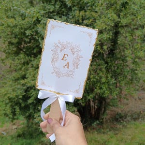 Puede incluir: Un abanico de boda blanco con bordes de purpurina dorada y un diseño floral dorado con las iniciales "E A" en el centro. El abanico tiene una cinta de satén blanca atada alrededor del mango.
