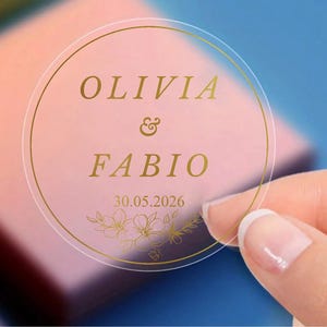Può includere: Un adesivo rotondo trasparente con scritte dorate e un bordo dorato. Il testo recita "OLIVIA & FABIO" con la data "30.05.2026" sotto. Design floreale in basso. L'adesivo è tenuto in mano.