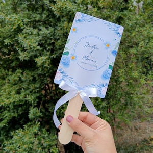 Può includere: Un ventaglio da matrimonio bianco con disegni floreali blu e bianchi. Il ventaglio ha un nastro bianco legato attorno al manico e dice "Jonathan & Mariana January 25, 2025".