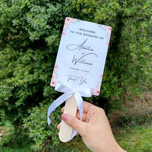 Puede incluir: Un abanico de boda blanco con borde floral rosa. El abanico dice "Bienvenido a la boda de Amelia & William, 25 de enero de 2026, Gracias".
