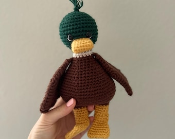 May include: A brown and green crocheted duck toy with yellow feet. The duck has black eyes and a white beak.