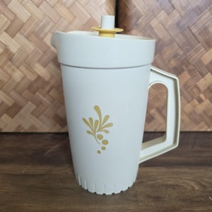 May include: A cream-colored plastic pitcher featuring a yellow floral graphic on the front. The pitcher has a sturdy handle, a matching lid with a small yellow knob, and a vertical ribbed design along the bottom edge of the container body.