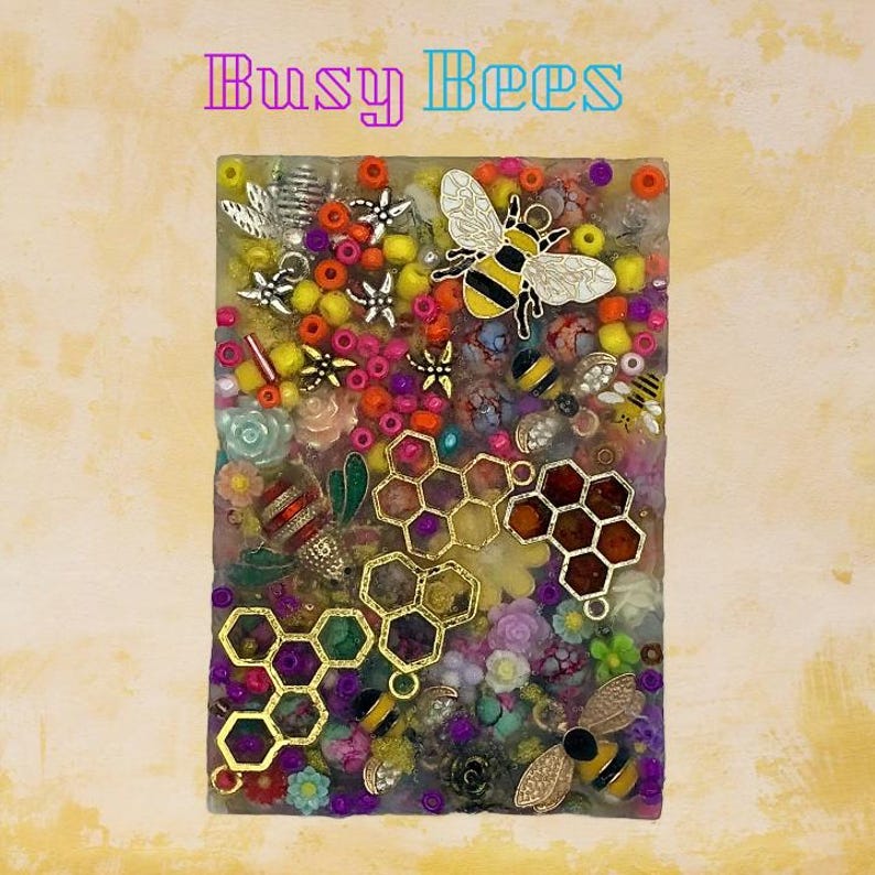 May include: A rectangular artwork with the text "Busy Bees" at the top. The artwork is filled with colorful beads, flowers, and bee-themed embellishments, including gold honeycomb shapes and bee charms. The background is a textured yellow.