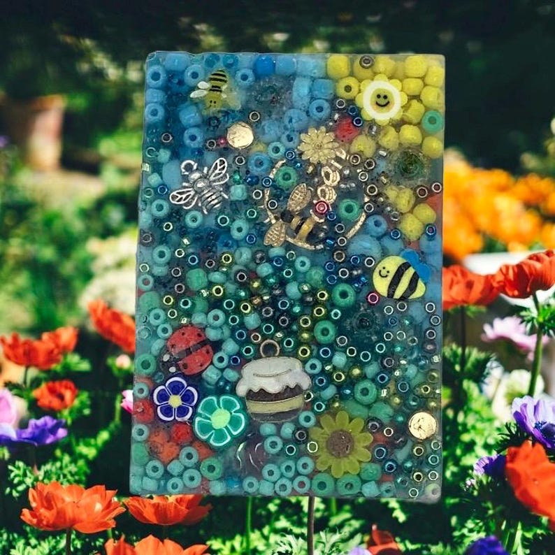 May include: A rectangular garden stake with a clear resin background filled with blue, green, and yellow beads. Decorative elements include bee, ladybug, and flower charms. The stake is placed in a garden with colorful flowers.