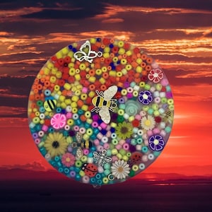 May include: A round, clear resin piece filled with colorful beads, flowers, and insect charms. The design includes a butterfly, bee, ladybug, and daisy. The background is a sunset.