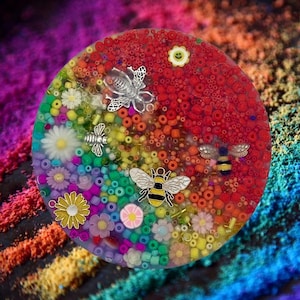May include: A round, colorful resin coaster with a rainbow of beads and flowers. Silver bee and flower charms are embedded in the resin. The coaster has a red section and a yellow smiling flower charm.