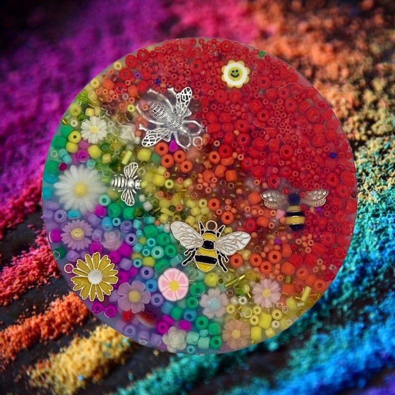 May include: A round, colorful resin coaster with a rainbow of beads and flowers. Silver bee and flower charms are embedded in the resin. The coaster has a red section and a yellow smiling flower charm.