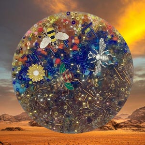 May include: A round resin coaster with a colorful mix of beads and metallic accents. The coaster features three bee charms and a yellow daisy charm. The background is a desert landscape with a sunset.