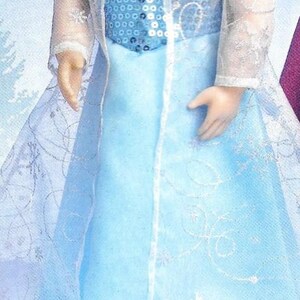 Peut inclure: Poup&eacute;e v&ecirc;tue d'une robe bleu clair avec un corsage &agrave; paillettes et une superposition transparente &agrave; motifs de flocons de neige. La poup&eacute;e a les cheveux blonds et porte une veste transparente &agrave; manches longues. La robe a une longue jupe fluide.
