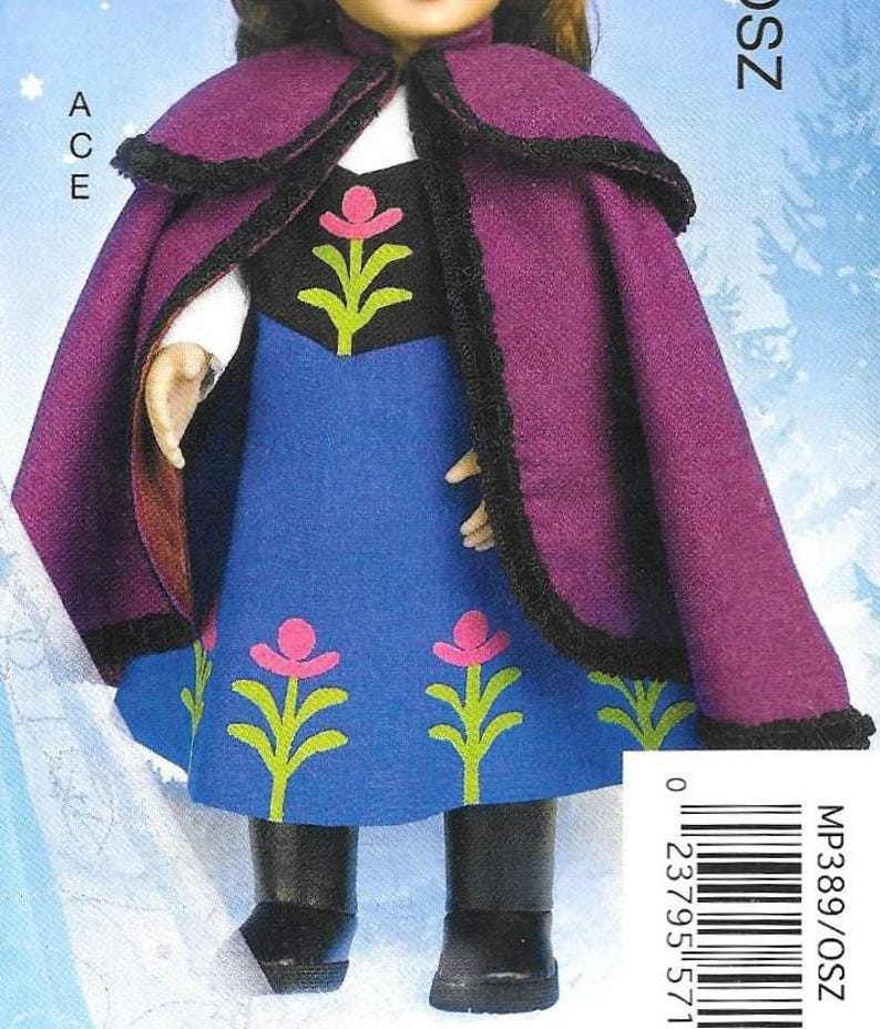 Peut inclure: Une poup&eacute;e v&ecirc;tue d'une cape violette avec une bordure noire et d'une robe bleue avec des motifs floraux roses et verts. La poup&eacute;e porte des bottes noires. L'image comprend le texte "ACE" et un code-barres.