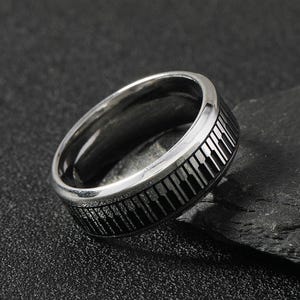 May include: A silver ring featuring a black piano key design. The ring is made from stainless steel and has a polished finish.