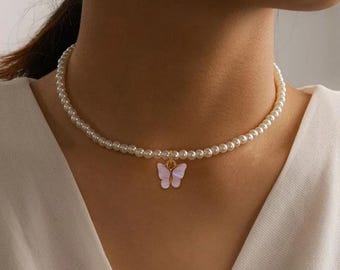 May include: A delicate pearl necklace with a small, light pink butterfly pendant. The butterfly has gold-colored accents. The necklace is worn around the neck, and the background is a white top.