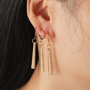 May include: Gold-coloured earrings with a dangling, tapered design. The earrings are shown on an ear, with three visible: two with a hook closure and one with a lever-back closure. The earrings have a polished finish and a simple, elegant style.