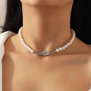 May include: A pearl necklace with a silver pendant. The necklace is made of white, round pearls. The pendant is silver-coloured and has a crown-like design. The necklace is worn around the neck, resting on the collarbone.