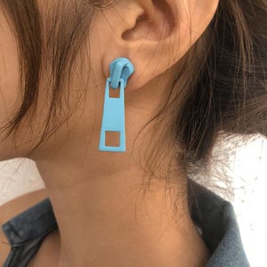 May include: A pair of light blue zipper earrings. The earrings are made to look like a zipper pull.
