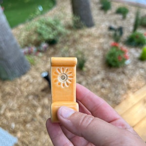 Peut inclure: Un crochet en c&eacute;ramique avec un motif floral de couleur orange clair. Le crochet a un petit trou au centre du motif.
