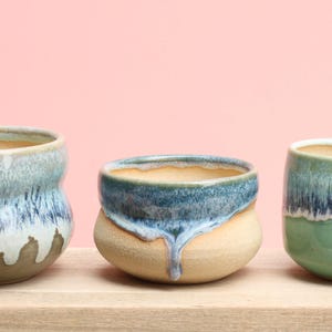 May include: Three ceramic pots with a unique glaze effect. The pots are in shades of blue, green, and tan, with a dripped glaze design. They sit on a wooden surface against a pink background. These pots are ideal for small plants.
