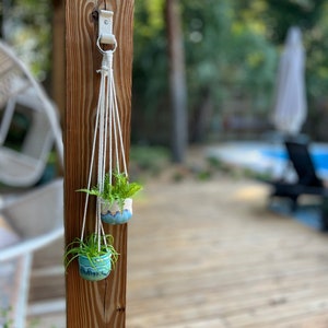 Peut inclure: Deux petits pots en c&eacute;ramique bleu et blanc avec des plantes vertes suspendus &agrave; un porte-plantes en macram&eacute; blanc fix&eacute; &agrave; un poteau en bois.