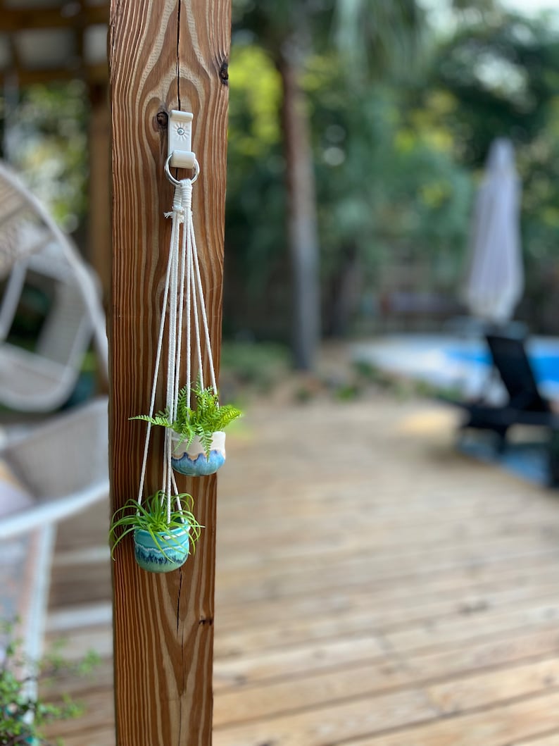 Peut inclure: Deux petits pots en c&eacute;ramique bleu et blanc avec des plantes vertes suspendus &agrave; un porte-plantes en macram&eacute; blanc fix&eacute; &agrave; un poteau en bois.