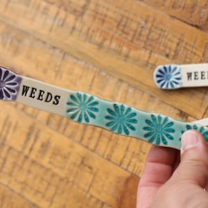 May include: A ceramic garden marker with a blue and green floral design. The marker is labeled "WEEDS".