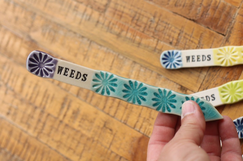 May include: A ceramic garden marker with a blue and green floral design. The marker is labeled "WEEDS".