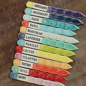 May include: A set of 12 ceramic plant markers with colorful glazes and white daisy-shaped patterns. Each marker is labeled with a different herb name: oregano, rosemary, sage, basil, marjoram, lavender, parsley, chives, cilantro, thyme, dill, and mint.