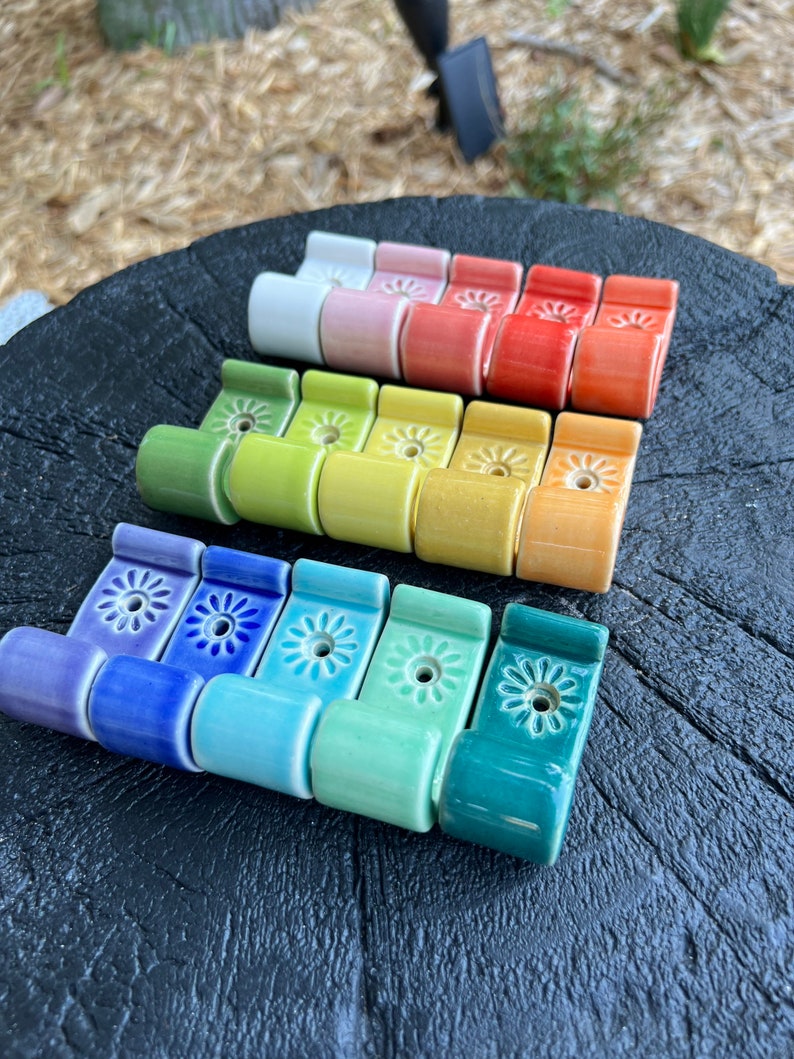 Peut inclure: Un ensemble de 12 crochets en c&eacute;ramique dans un arc-en-ciel de couleurs, chacun avec un petit motif de fleur. Les crochets sont dispos&eacute;s en rang&eacute;e, en commen&ccedil;ant par le blanc et en terminant par le bleu sarcelle.
