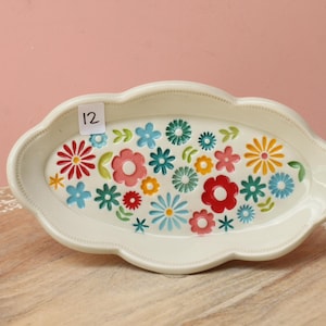 May include: An oval-shaped cream-colored ceramic tray with a scalloped edge and a beaded rim. The interior surface features a colorful pattern of various small flowers in shades of red, yellow, blue, and green. A small white sticker with the number 12 is attached.