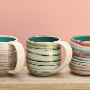 May include: Three ceramic mugs with white handles and teal interiors. Each mug features a unique horizontal striped pattern in shades of blue, green, and red. The mugs are displayed on a wooden surface against a pink background.