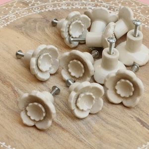 May include: A collection of off-white ceramic flower-shaped cabinet knobs with silver screws. The knobs are arranged on a light brown wooden tray with a decorative border. The knobs are approximately 1.5 inches in diameter.