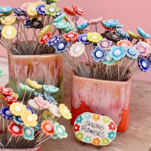 May include: Colorful ceramic flower garden stakes in various colors, including blue, yellow, red, and white, are displayed in square ceramic pots. A sign that reads "Spring Flowers" is also present. The stakes are on a wooden surface.