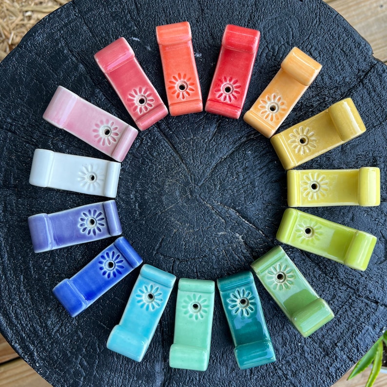 Peut inclure: Un ensemble de 13 crochets en c&eacute;ramique de diff&eacute;rentes couleurs, chacun avec un motif de fleur en relief. Les crochets sont dispos&eacute;s en cercle sur une surface en bois fonc&eacute;.