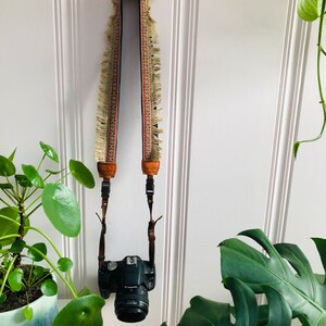 Peut inclure: Un appareil photo noir avec une sangle en cuir marron accroch&eacute; &agrave; un mur blanc. La sangle est orn&eacute;e d'une frange et d'un motif g&eacute;om&eacute;trique. L'appareil photo est accroch&eacute; &agrave; un crochet vert.