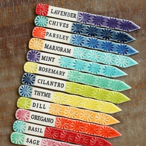 May include: Set of 12 colorful ceramic plant markers with flower designs and names of herbs written in black. The markers are arranged in a rainbow pattern from purple to pink.