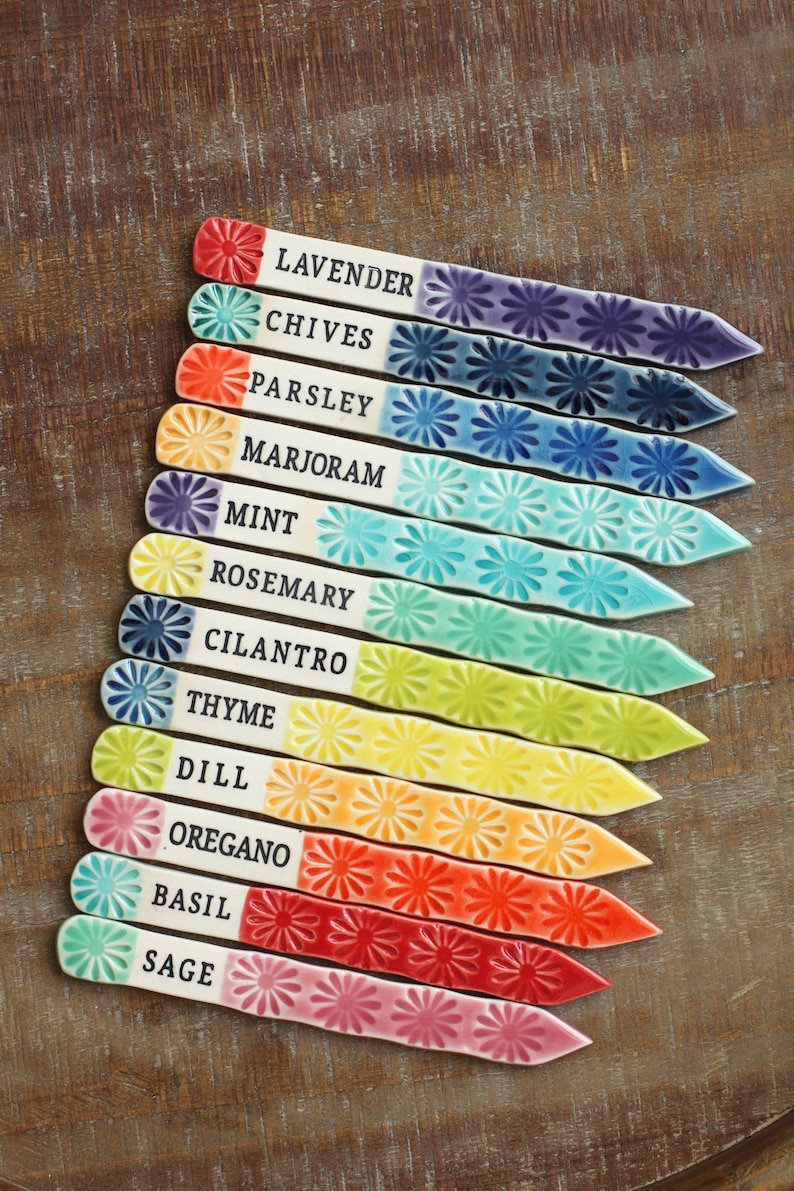 May include: Set of 12 colorful ceramic plant markers with flower designs and names of herbs written in black. The markers are arranged in a rainbow pattern from purple to pink.