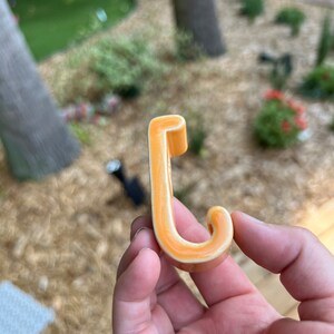 Peut inclure: Un crochet en c&eacute;ramique en forme de lettre J. Le crochet est de couleur orange clair avec un contour blanc.
