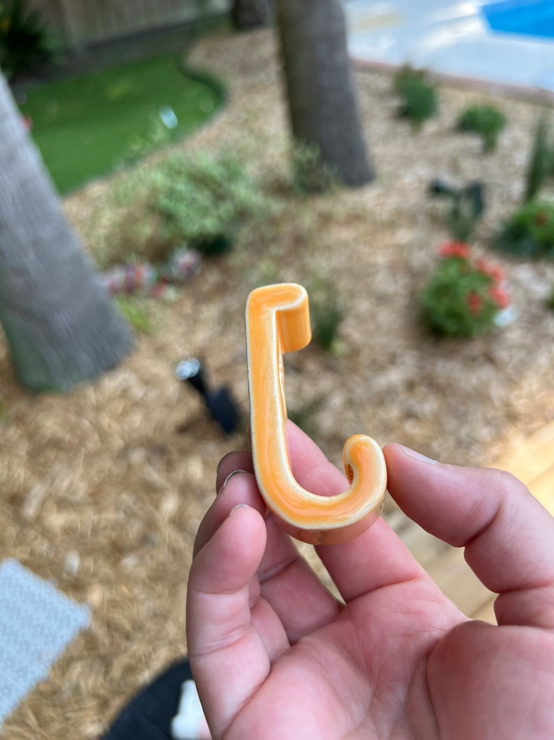 Peut inclure: Un crochet en c&eacute;ramique en forme de lettre J. Le crochet est de couleur orange clair avec un contour blanc.