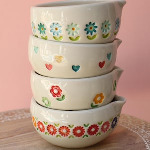 May include: A stack of four cream-colored ceramic bowls with pouring spouts. The top bowl features blue and green flowers, the second has small colorful hearts, the third displays red and orange flowers, and the bottom bowl has a band of red flowers.