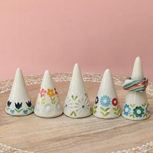 May include: Five white ceramic cone-shaped ring holders arranged in a row. Four holders feature painted floral patterns in blue, yellow, red, and green. One holder on the far right displays a textured white floral design and holds a colorful striped ring.