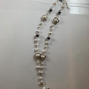 Puede incluir: Un collar de perlas blancas con dijes de coraz&oacute;n plateados y un dije de &aacute;ngel. El collar est&aacute; sobre un fondo blanco.