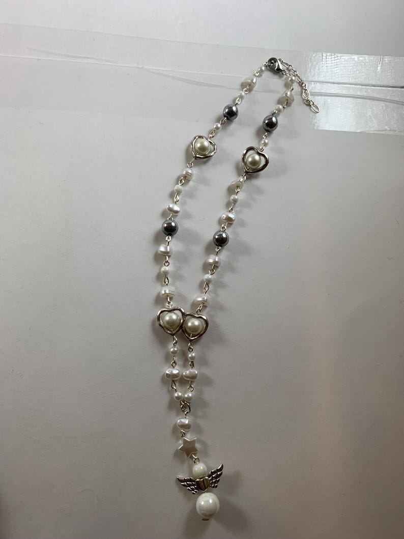 Puede incluir: Un collar de perlas blancas con dijes de coraz&oacute;n plateados y un dije de &aacute;ngel. El collar est&aacute; sobre un fondo blanco.