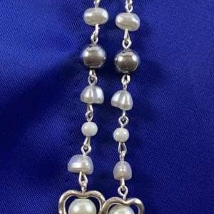 Puede incluir: Un collar de cadena de plata con cuentas de perlas blancas y dijes de coraz&oacute;n de plata. El collar tiene un dije de &aacute;ngel de plata con una estrella y una cuenta de perla en la parte inferior.