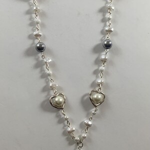 Puede incluir: Un collar de perlas blancas con dijes de coraz&oacute;n plateados y un dije de &aacute;ngel plateado con una estrella en la parte superior. El collar tiene un cierre plateado.