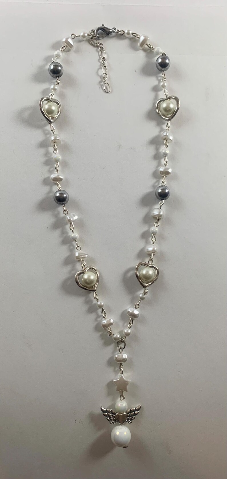 Puede incluir: Un collar de perlas blancas con dijes de coraz&oacute;n plateados y un dije de &aacute;ngel plateado con una estrella en la parte superior. El collar tiene un cierre plateado.