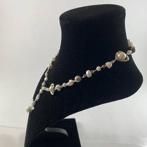 Puede incluir: Un collar de plata con perlas blancas y grises y un colgante en forma de coraz&oacute;n. El collar se muestra en un soporte de terciopelo negro.